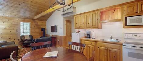 L’intérieur d’une cabane en bois comprend une table à manger, des chaises, une cuisine avec des meubles en bois, un réfrigérateur, un four à micro-ondes et un ventilateur de plafond.
