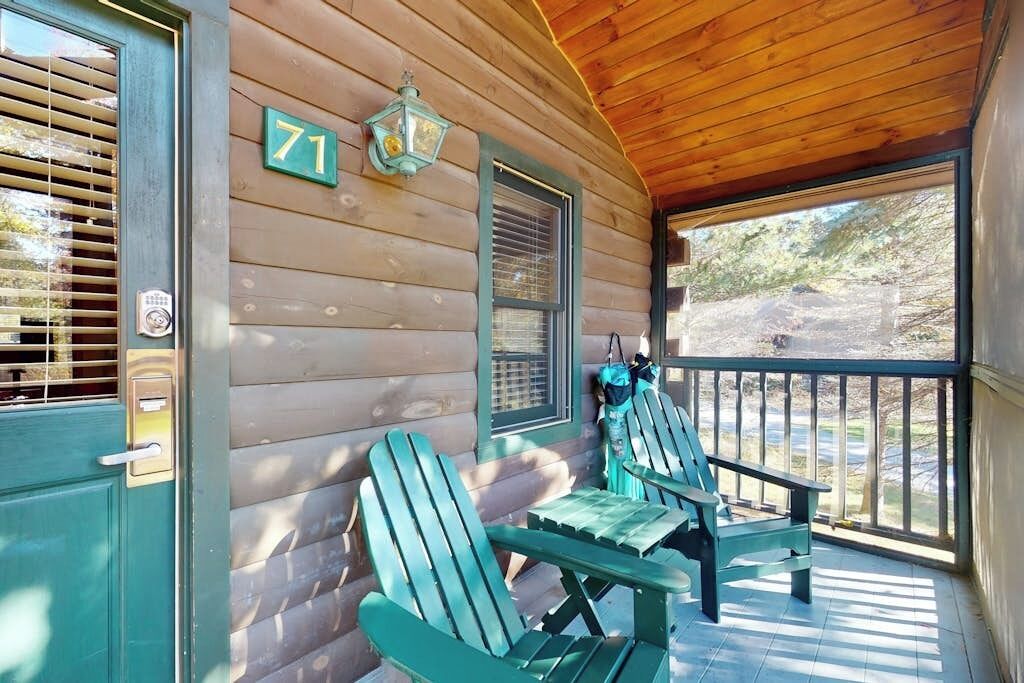 A wooden cabin with a porch, two Adirondack chairs, a number '71' sign, and a window with shutters.
