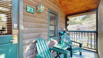 A wooden cabin with a porch, two Adirondack chairs, a number '71' sign, and a window with shutters.