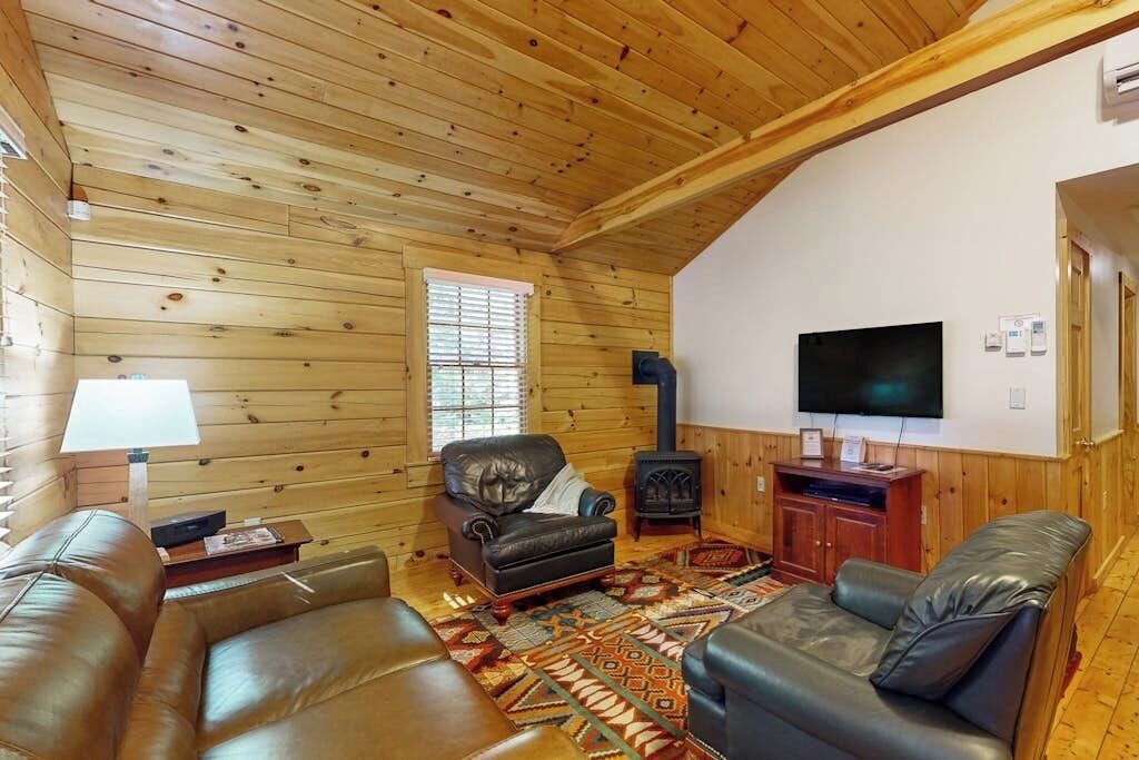 A log cabin interior with a fireplace, leather sofas, a television, and a patterned rug.