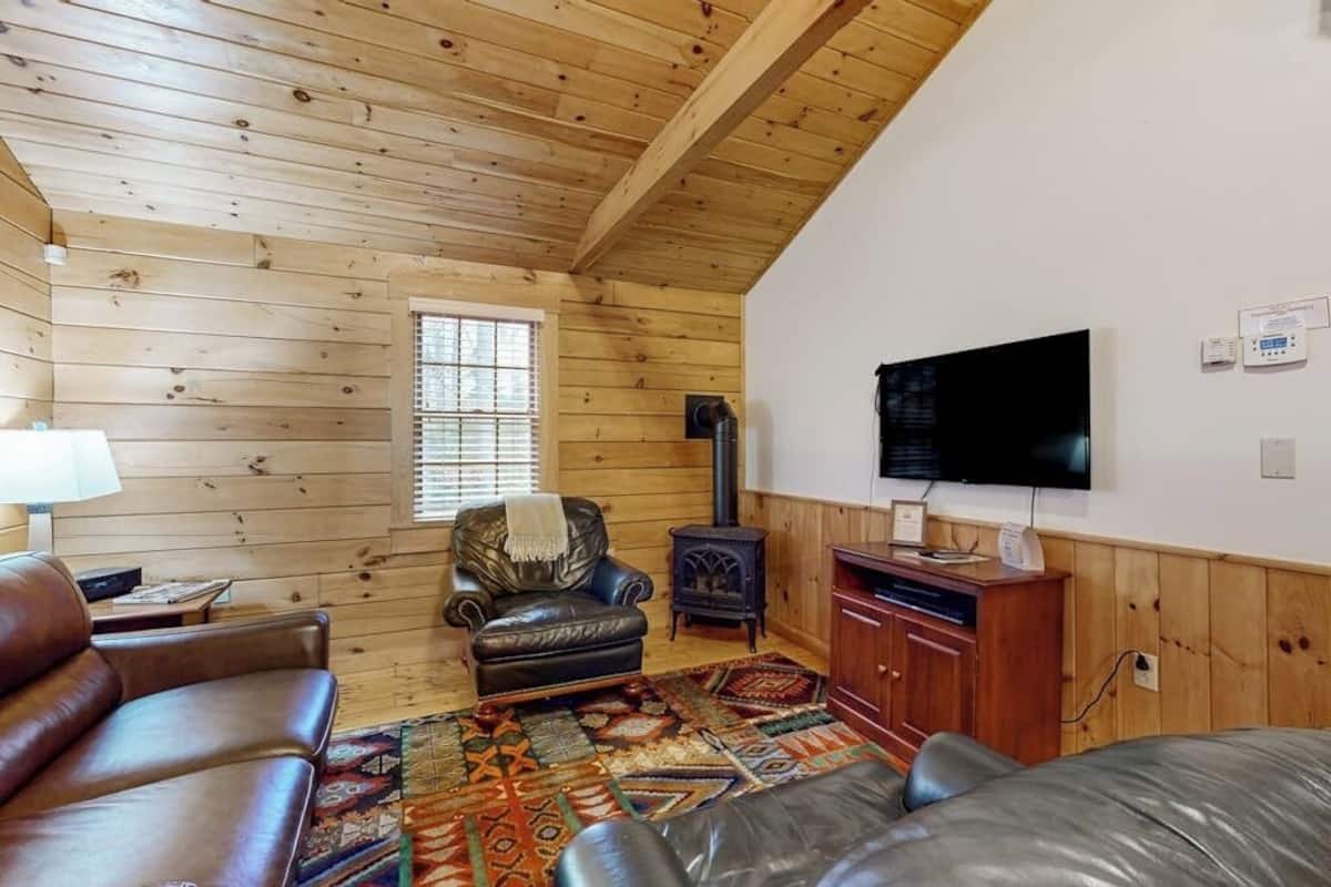 A cozy cabin interior with a wooden ceiling, a fireplace, a television, and leather furniture.