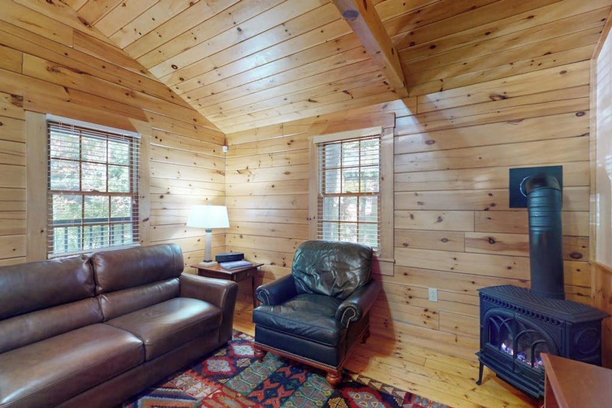 A log cabin interior with a leather sofa, armchair, and wood-burning stove.