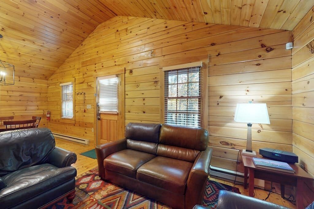A log cabin interior with a leather sofa, a small table with a lamp, and a fireplace.