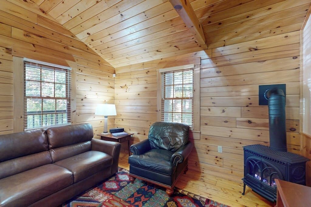 A log cabin interior with a leather sofa, armchair, and wood-burning stove.