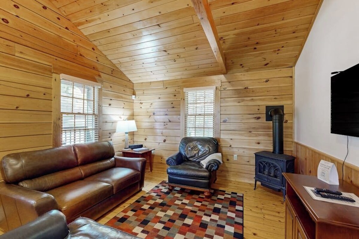 Interior de una cabaña de madera con sofá, sillón, alfombra, chimenea y televisor.