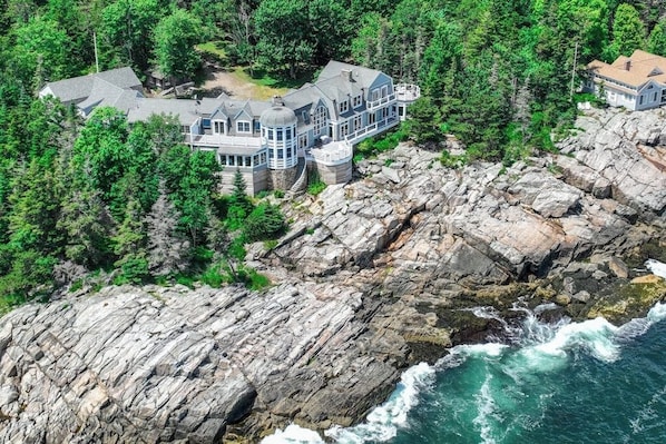 A large, multi-story house with a stone foundation, surrounded by dense forest and rocky coastline. - Spectacular Oceanfront Retreat With Amazing Views, Sunroom & Washer/dryer (Georgetown)