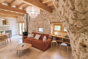 Ein rustikales Interieur mit Steinwand, Holzbalken, einer Couch, einem Esstisch und einer Küchenzeile.
