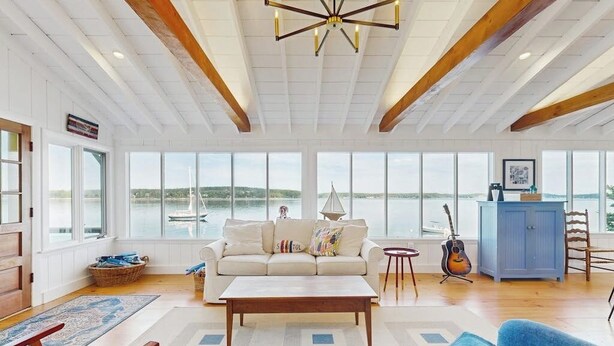 A spacious living room with a large window overlooking a body of water, a white sofa, a coffee table, and a guitar. - Charming Cottage: Deepwater Dock, Panoramic Linekin Bay Views, Near Village (Boothbay)