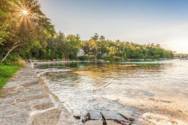A stone pathway along a riverbank with a view of a forested area and buildings in the background. - Amazing All-season Sprucewold Cabin w/ Private Beach Club Access Dog-friendly (Boothbay Harbor)