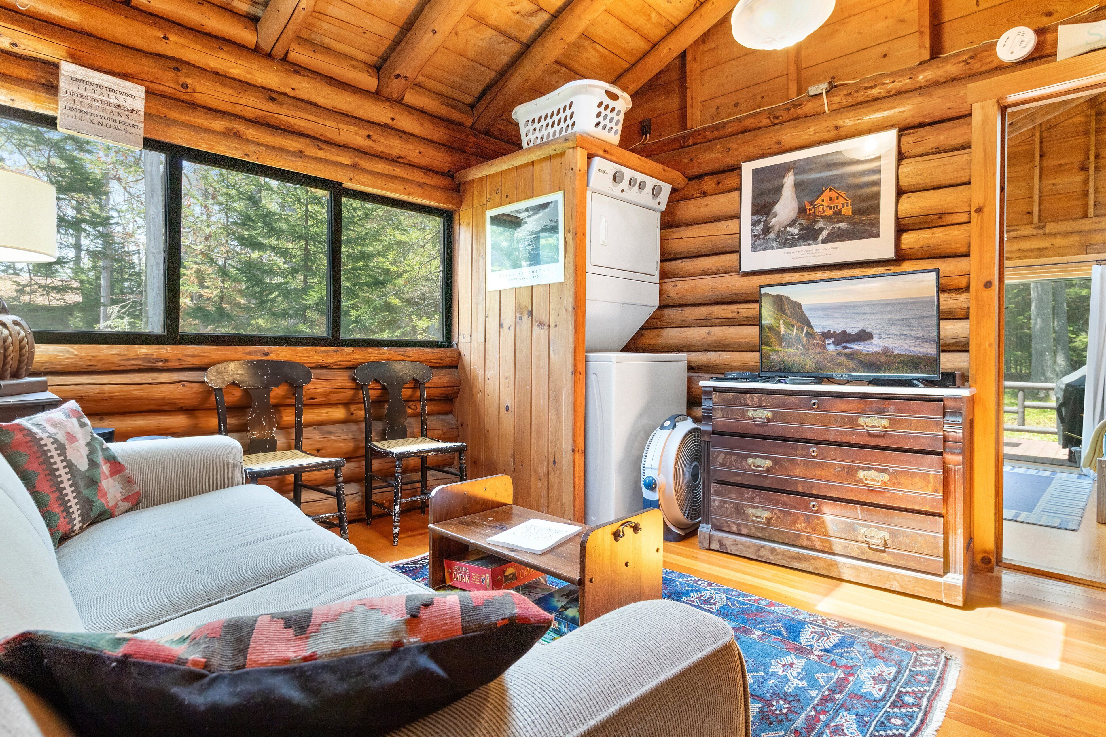 A log cabin interior with a sofa, chairs, a table, a dresser, a washing machine, and a framed picture on the wall.