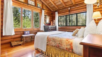 Dormitorio de cabaña de madera con cama, cómoda y ventanas con vistas a un bosque.