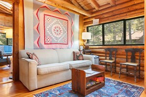 A log cabin interior with a sofa, wooden chairs, a quilt, and a view through large windows. - Cozy Home With Serene Views, Wood-burning Fireplace & Wifi (Boothbay Harbor)