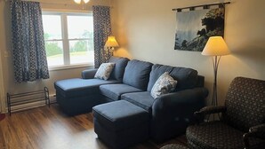 Een woonkamer met een hoekbank, voetenbankje en gestreept fauteuil. Er is een plafondventilator, een raam met gordijnen en een schilderij aan de muur.