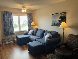 Uma sala de estar com sofá seccional, um banquinho e uma poltrona estampada. Há um ventilador de teto, uma janela com cortinas e um quadro na parede.