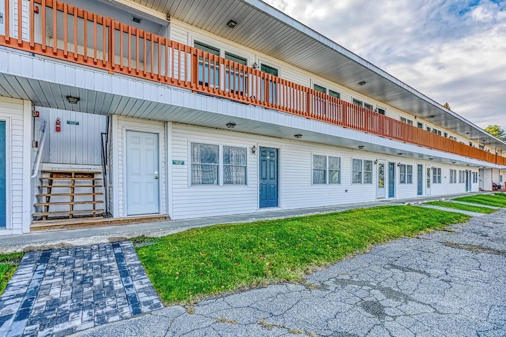 A row of white motel buildings with orange railings, a paved walkway, and a grassy area.