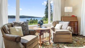 A room with a large window offering a view of a lake and forest, two leather armchairs, a wooden side table with a fan, and a dresser with a lamp. - Dog-friendly Oceanfront Home With Private Shorefront, two Fireplaces, & Firepit (Steuben)