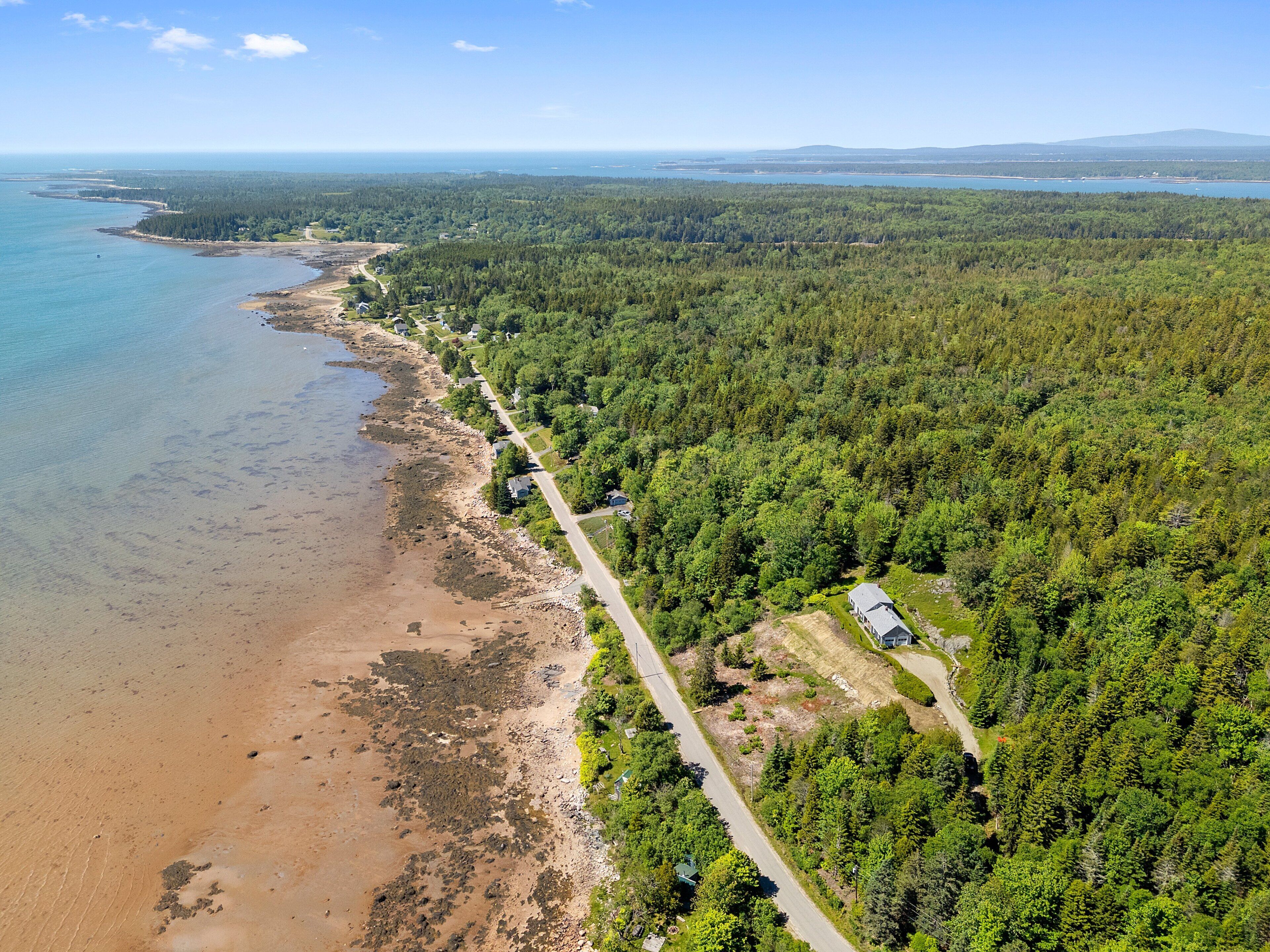 An aerial view of a coastal area with a road running alongside a beach, surrounded by dense forest.