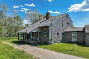 A rustic wooden house with a porch, a chimney, and a flag. - Waterfront Stay With Scenic Deck, Grill, Smart Tvs, Shore Access - Dog-friendly (Penobscot)