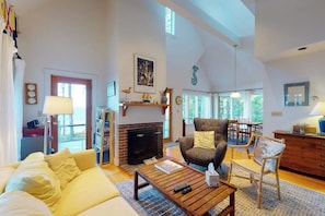 A spacious living room with a fireplace, a yellow sofa, a wooden coffee table, and a large window. - Dog-friendly, Waterfront Home on St. George River With Shore Access, Great Views (Port Clyde)