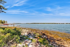 A rocky shoreline with clear water, a dock, and distant trees. - Dog-friendly, Waterfront Home on St. George River With Shore Access, Great Views (Port Clyde)