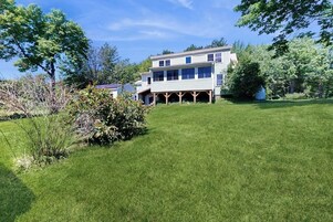 Uma casa de dois andares com varanda coberta, cercada por um gramado bem cuidado e árvores.