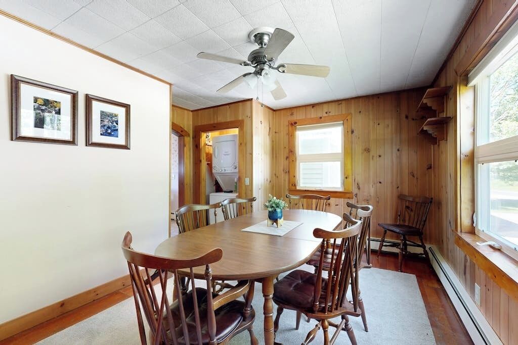 A wooden-paneled room with a dining table, chairs, ceiling fan, framed pictures, and a window.