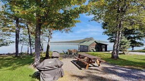 A lakeside cabin with a picnic table and grill, surrounded by trees and a view of the lake. - Dog-friendly Lakefront Cabin With Private Dock, Epic Views of Lake & Mt Kineo (Rockwood)