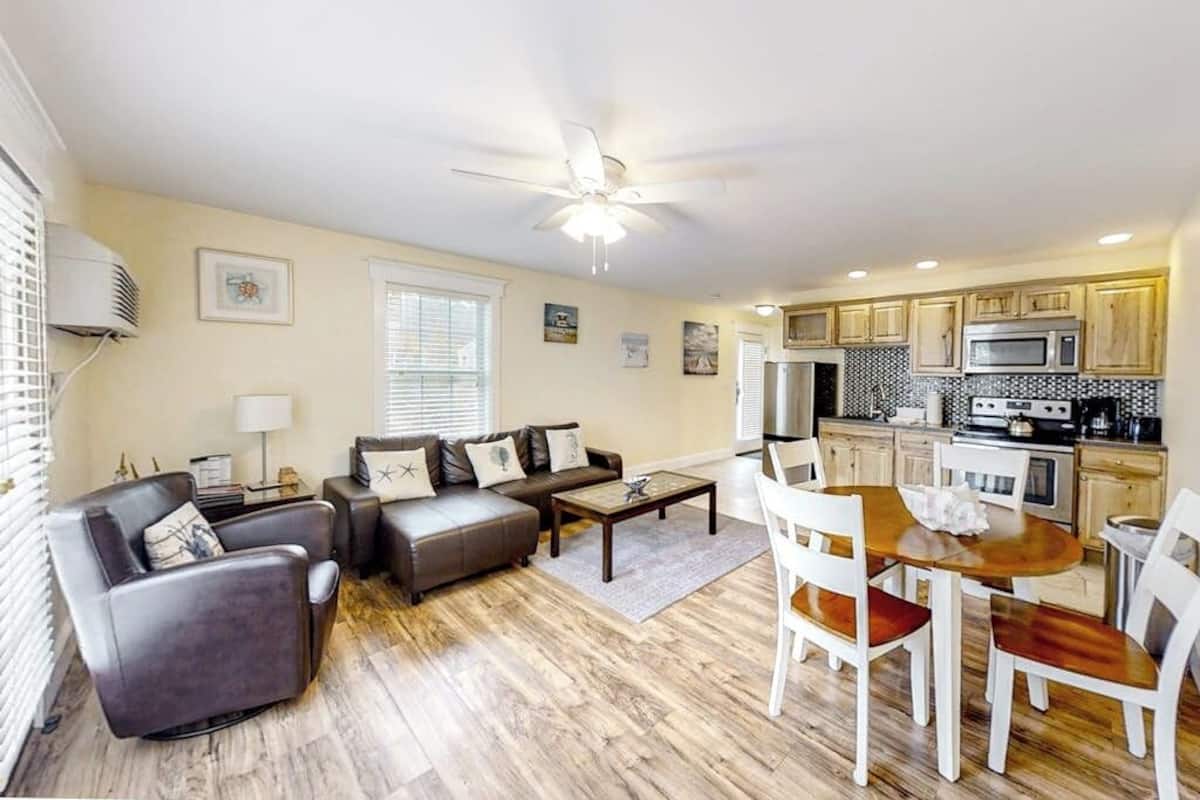 A living room with a sofa, chairs, a coffee table, and a dining table with chairs. There is a kitchen area with cabinets and appliances.