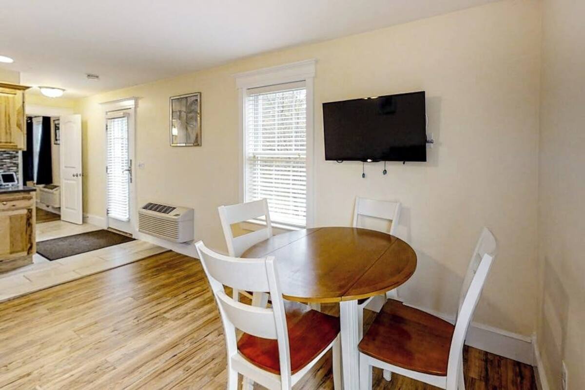 A dining area with a round table, white chairs, a flat-screen TV mounted on the wall, and a kitchen in the background.