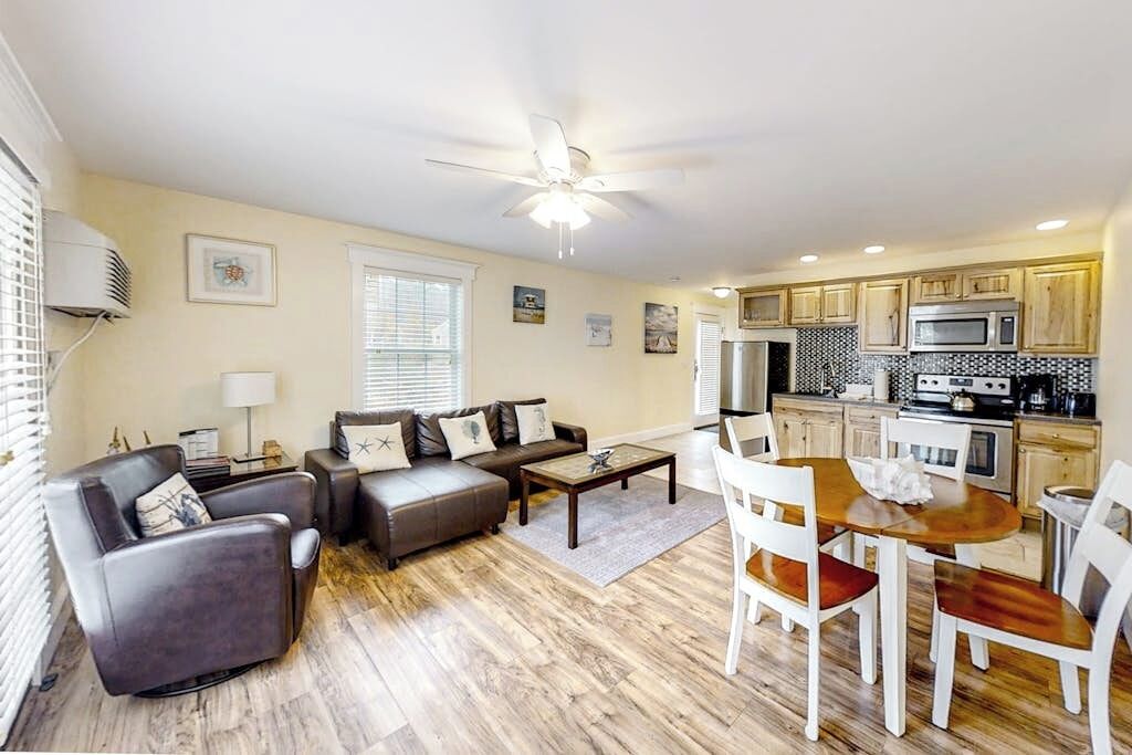 A living room with a sofa, chairs, a coffee table, and a dining table with chairs. There is a kitchen area with cabinets and appliances.