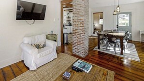 Un soggiorno con un divano bianco, una TV a schermo piatto, un pilastro in mattoni, un'area pranzo con tavolo e sedie, e una cucina sullo sfondo.