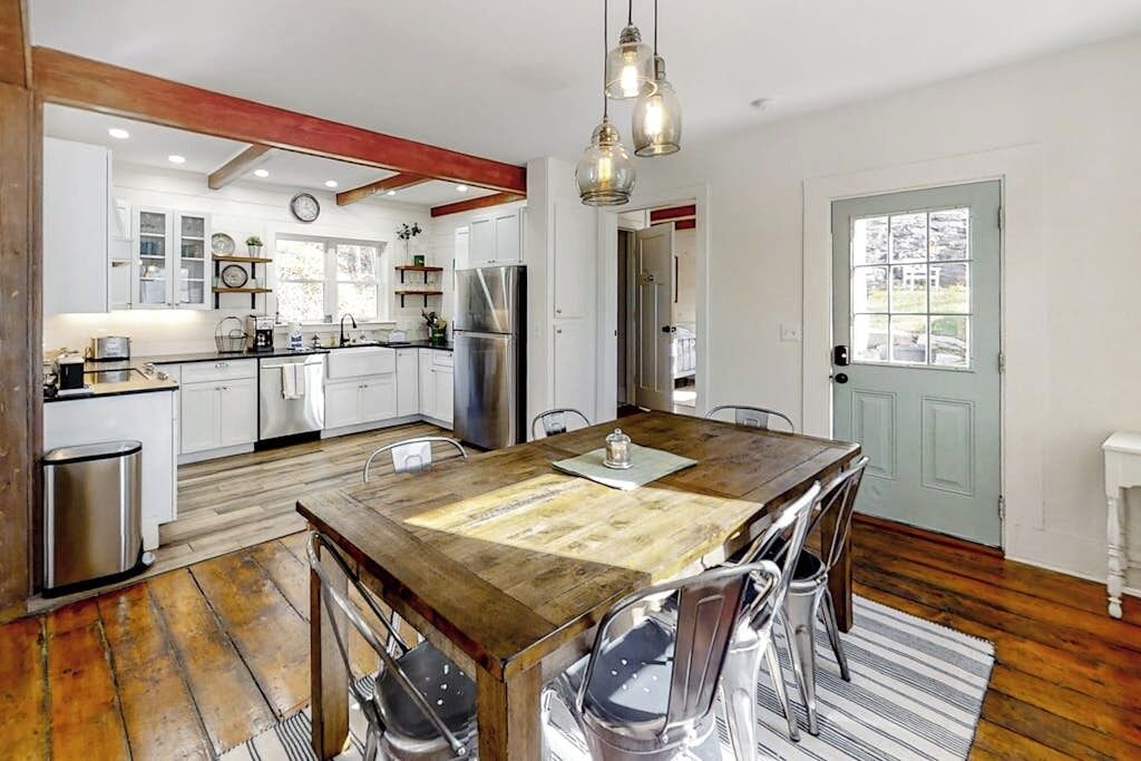 A kitchen with white cabinets, stainless steel appliances, a wooden dining table, and a hanging light fixture.
