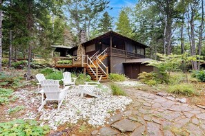 Eine Hütte mit einer Terrasse, umgeben von Bäumen, und zwei Adirondack-Stühlen auf einem Steinweg.