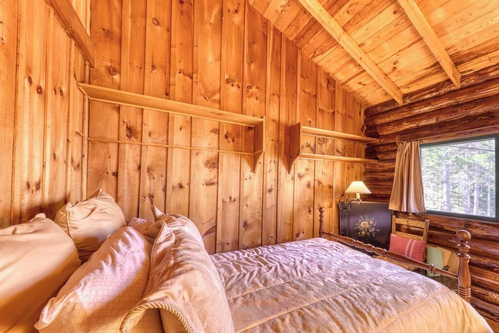 A wooden cabin bedroom with a bed, pillows, a nightstand, a lamp, and a window.