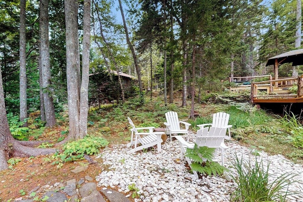 A forested area with a wooden deck, white Adirondack chairs, and a rustic cabin.