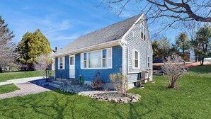 A single-story house with a gabled roof, a front porch, and a small front yard with a pathway leading to the entrance. - Charming Coastal House With Cozy Living Spaces and Outdoor Firepit (Winter Harbor)