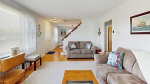 A living room with a sofa, a coffee table, a staircase, and a painting on the wall. - Charming Coastal House With Cozy Living Spaces and Outdoor Firepit (Winter Harbor)
