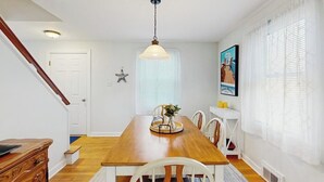 A dining area with a wooden table, white chairs, a hanging light fixture, and a staircase. - Charming Coastal House With Cozy Living Spaces and Outdoor Firepit (Winter Harbor)