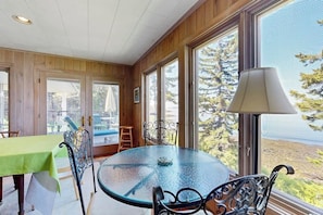A room with a round glass table, metal chairs, a green tablecloth, and large windows with a view of trees and a body of water. - Beachfront Delight With Detached Guesthouse, Private Beach Access, Dog-friendly (Southport)