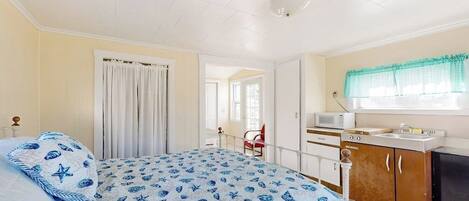 Un dormitorio con cama, kitchenette, ventana con cortinas y puerta que da a otra habitación.