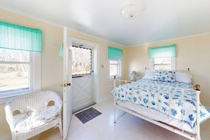 A bedroom with a bed, a chair, a door, and windows with curtains. - Charming Love Shack With Stunning Seaside Views and all the Comforts of Home (Trenton)