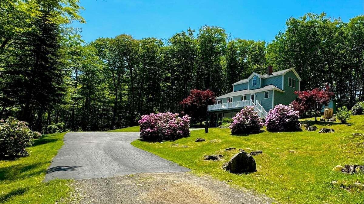 A two-story house with a driveway leading to the front door, surrounded by lush greenery and flowering bushes.