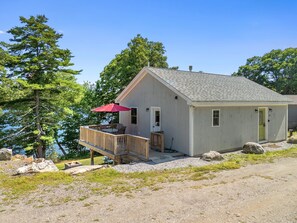 A small cabin with a deck, a red umbrella, and a view of the water. - Cozy Lakefront Cottage With Stunning Views, Deck, BBQ Grill, and Outdoor Firepit (Newcastle)