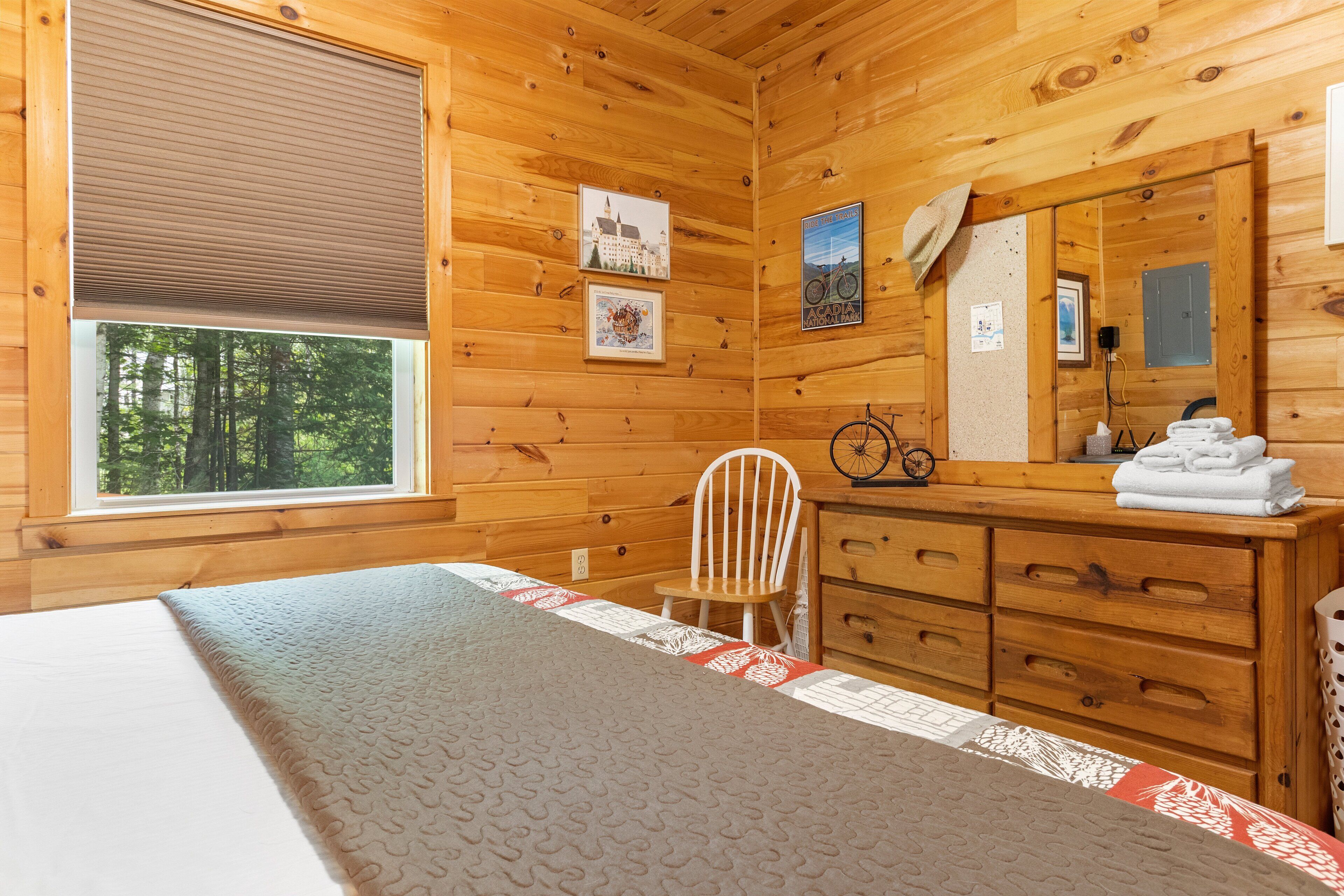 A wooden cabin room with a bed, a chair, a dresser, and a window with a view of trees.