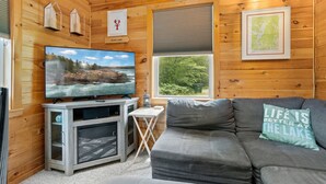 A wooden cabin interior with a television, a gray sofa, and a window with a view of trees. - Waterfront Home on Graham Lake With Kayaks, Bikes, Firepit & Fireplace (Ellsworth)