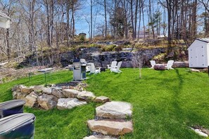 Ein Garten mit Rasenfläche, weißen Stühlen, einem Schuppen und einer Steinmauer.