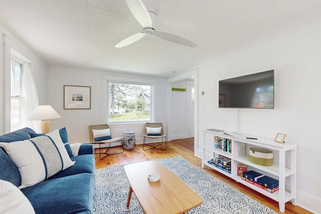 A living room with a blue sofa, a wooden coffee table, a white bookshelf, a ceiling fan, and a TV mounted on the wall.