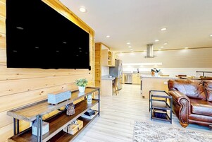 Uma sala de estar moderna com uma TV grande, prateleiras de madeira, um sofá de couro e uma cozinha ao fundo.