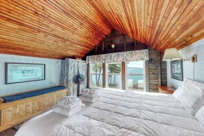 A bedroom with a wooden ceiling, a large bed, a bench, a fan, and a window with a view. - Spacious Waterfront Estate & Cabins W/ Porch, Fireplace, Kayaks & Free Wifi (Brunswick)