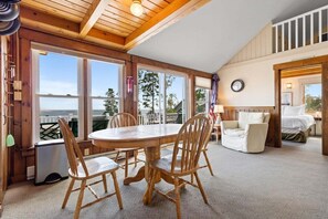 A cozy room with a wooden dining table and chairs, a bed, and large windows offering a view. - Little White Caps, Quaint Maine Cottage (Tenants Harbor)